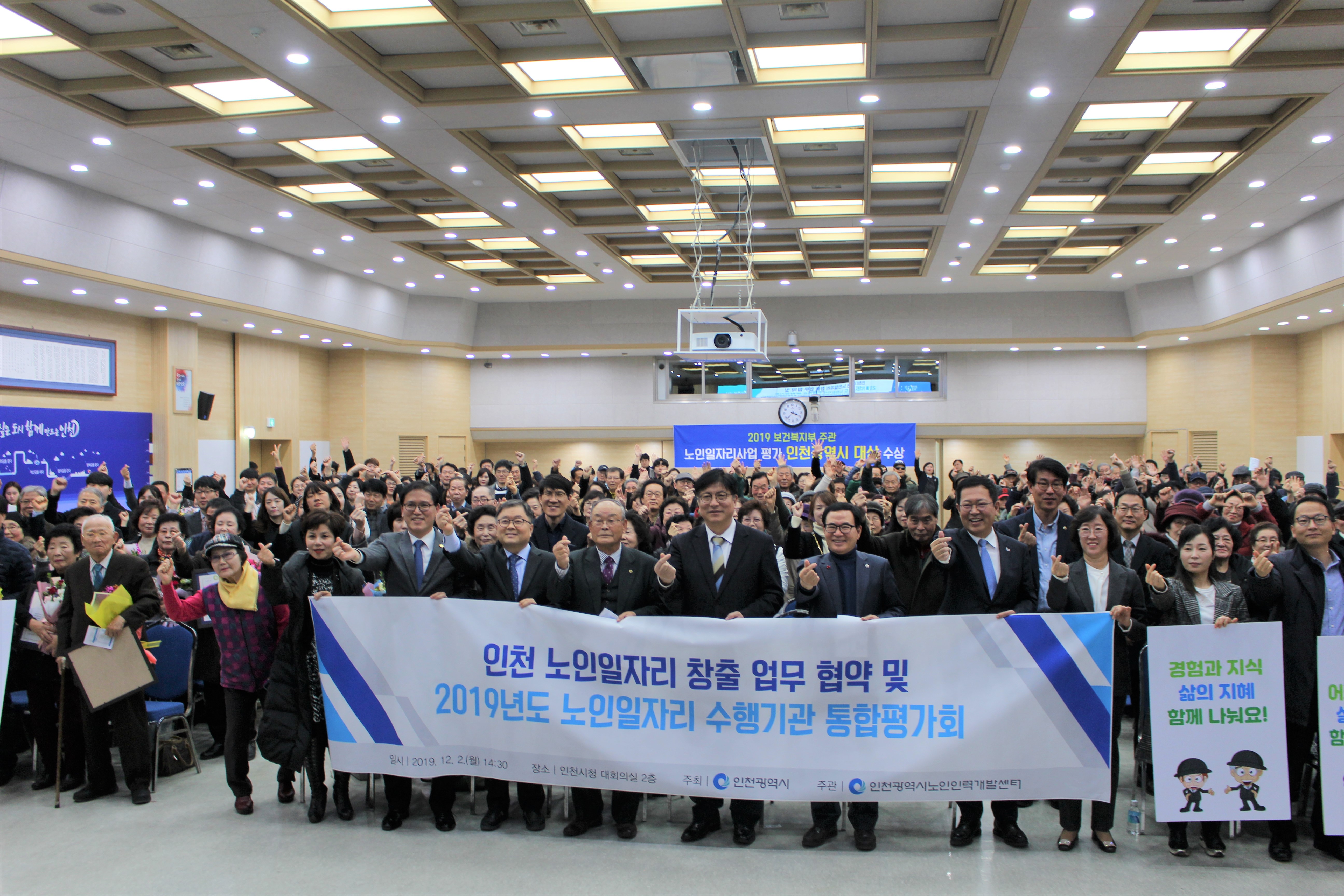 강당에 가득한 사람들이 정면을 바라보고 서있으며, 맨 앞줄의 사람들이 