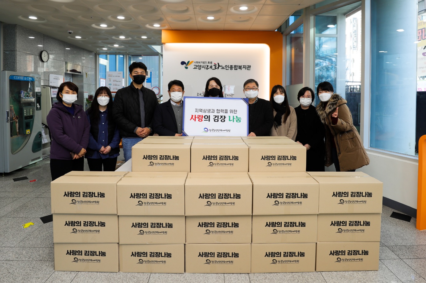 다수의 사람들이 정면을 바라보며 서 있고, 그 앞에 많은 박스가 쌓여있는 모습